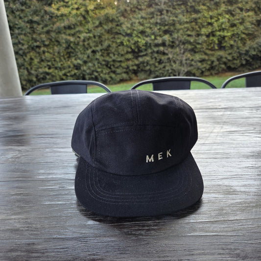 Gorra MEKap Five Panel en negro, de frente, con el wordmark MEK bordado en hilo dorado